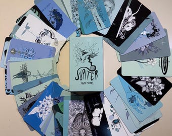 May include: A deck of tarot cards arranged in a circular fan pattern around a central box labeled JUPITER POWER TAROT. The cards feature various black and white line art illustrations of faces, hands, and flowers on light blue and black backgrounds.