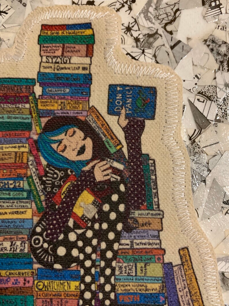 May include: A woman with blue hair is surrounded by a stack of books. She is holding a book that says "Don't Panic!" The woman is wearing a black and white polka dot dress.