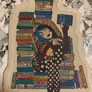 May include: A colourful fabric patch depicting a cartoon woman with blue hair sitting on a stack of books. The woman is holding a book that reads "Don't Panic". The books are stacked high and include titles such as "Catcher in the Rye", "The Hitchhiker's Guide to the Galaxy", and "The Lord of the Rings".
