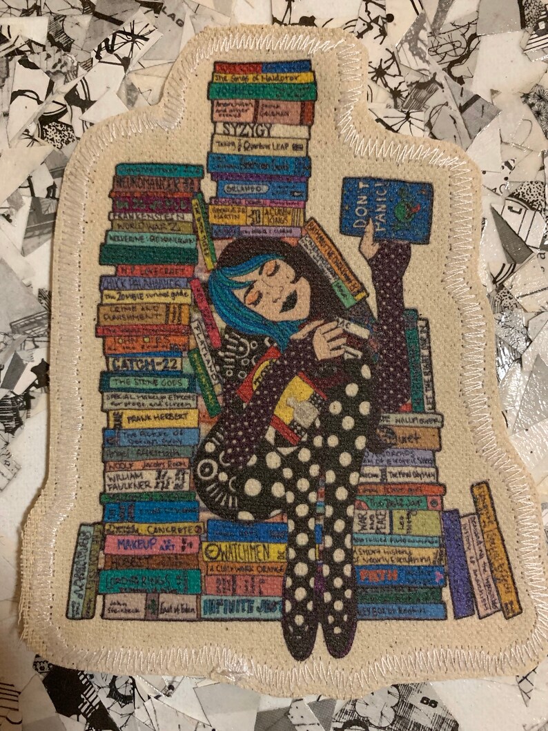 May include: A colourful fabric patch depicting a cartoon woman with blue hair sitting on a stack of books. The woman is holding a book that reads "Don't Panic". The books are stacked high and include titles such as "Catcher in the Rye", "The Hitchhiker's Guide to the Galaxy", and "The Lord of the Rings".