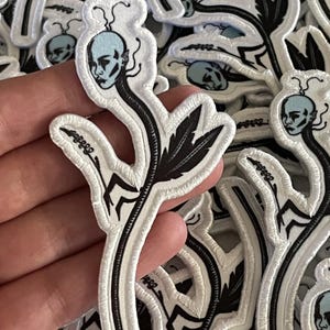 May include: A white embroidered patch with a black outline depicting a stylized human head with a blue face and black hair growing from the top of the head. The head is attached to a black stem with a black leaf. The patch is surrounded by other similar patches.