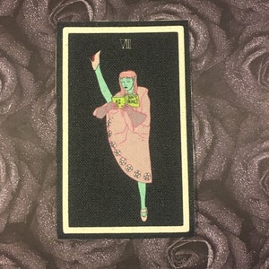 Peut inclure: Illustration de carte de tarot avec une figure à la peau verte et une robe rose, levant une jambe. La carte a un fond noir avec le chiffre romain VII. Un petit panneau indique "DON'T PANIC" et le numéro "42".