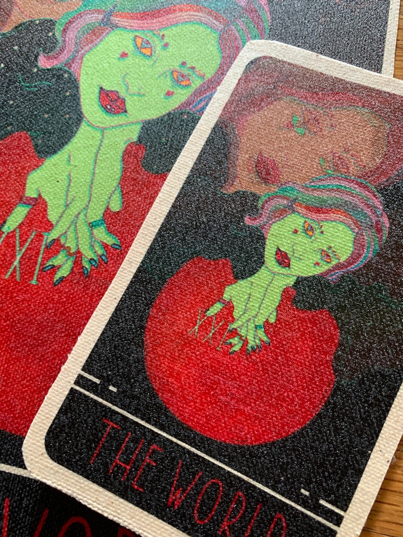 May include: Tarot card featuring a green-skinned figure with red lips and multi-colored hair. The figure is set against a red background with a black border. The text "THE WORLD" is visible at the bottom of the card.