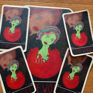 May include: A tarot card set featuring a central mat and four cards, all with a similar design. The artwork depicts a green-skinned figure with colorful hair and a red dress. The cards are labeled "THE WORLD" in white text.