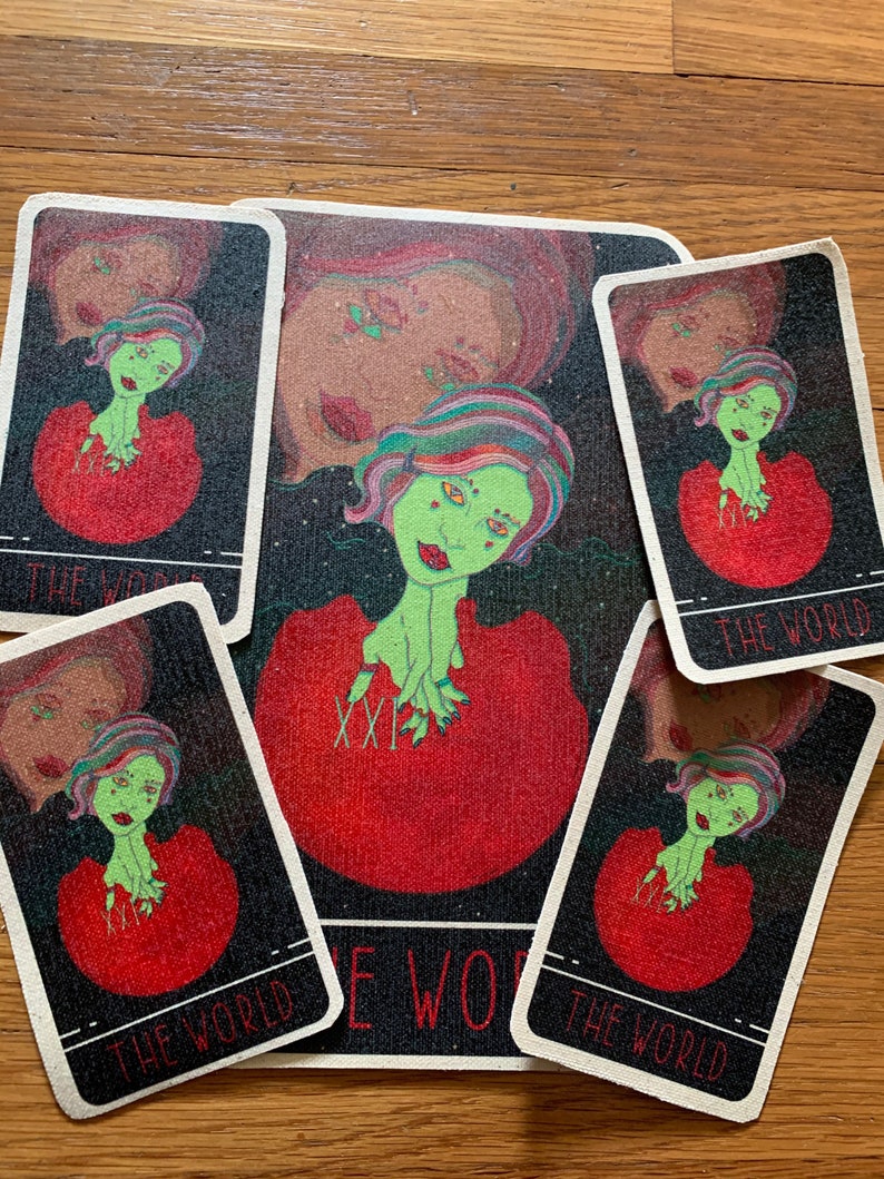 May include: A tarot card set featuring a central mat and four cards, all with a similar design. The artwork depicts a green-skinned figure with colorful hair and a red dress. The cards are labeled "THE WORLD" in white text.