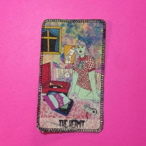 Può includere: Illustrazione di carta dei tarocchi con una figura in un vestito a pois rossi e bianchi, con pelle verde e capelli argentati. La carta raffigura una stanza con una finestra, un giradischi e il testo "THE HERMIT". La carta ha un bordo cucito nero.