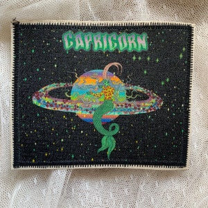 Op de afbeelding: Zwart rechthoekig embleem met het woord "CAPRICORN" in groen, met een kleurrijke illustratie van een zeemeerminachtig figuur met een planeet en ringontwerp. De achtergrond is een sterrennacht.