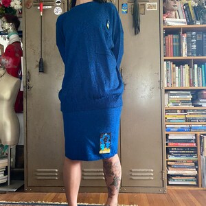 May include: A two-piece blue knit sweater set. The sweater has a pocket and a small yellow emblem on the chest. The skirt has a patch with a colorful design. The set is worn by a person standing in front of a metal locker.