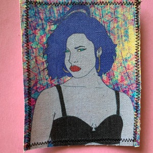 Puede incluir: Un fondo abstracto colorido con un rostro de mujer en el centro. Ella tiene el pelo azul, los ojos azules y lleva una blusa negra.