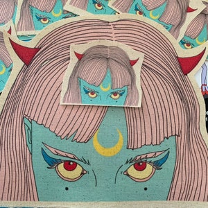 Può includere: Un'illustrazione blu e rosa di una donna con una luna crescente sulla fronte e corna rosse. I suoi occhi sono spalancati e ha un'espressione seria. L'illustrazione è su uno sfondo di tessuto beige.