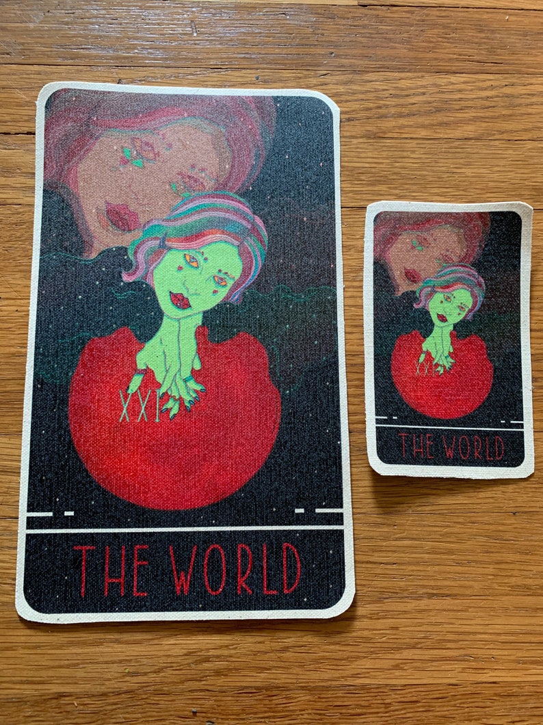 May include: Two tarot cards, one large and one small, featuring a green-skinned figure with colorful hair and a red dress. The cards have a black starry background and the text "THE WORLD" at the bottom. The larger card is approximately 4 x 6 inches.