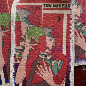 Könnte beinhalten: Rechteckiger Aufnäher mit Tarotkarten-Design. Das Bild zeigt zwei sich umarmende Figuren in Rot- und Grüntönen. Der Text "THE LOVERS" steht oben. Der Aufnäher hat einen weißen, genähten Rand.