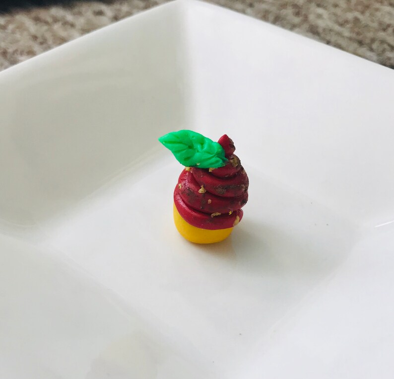 Pode incluir: Um pequeno cupcake vermelho e amarelo com uma folha verde no topo. O cupcake &eacute; decorado com granulado dourado.