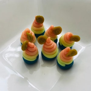 Pode incluir: Seis pequenos doces coloridos em forma de espiral com uma folha verde no topo. Os doces est&atilde;o dispostos em um prato branco.