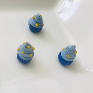 Pode incluir: Tr&ecirc;s mini cupcakes azuis e amarelos com cobertura branca. Os cupcakes s&atilde;o feitos de argila polim&eacute;rica e t&ecirc;m aproximadamente o tamanho de uma moeda de 10 c&ecirc;ntimos.