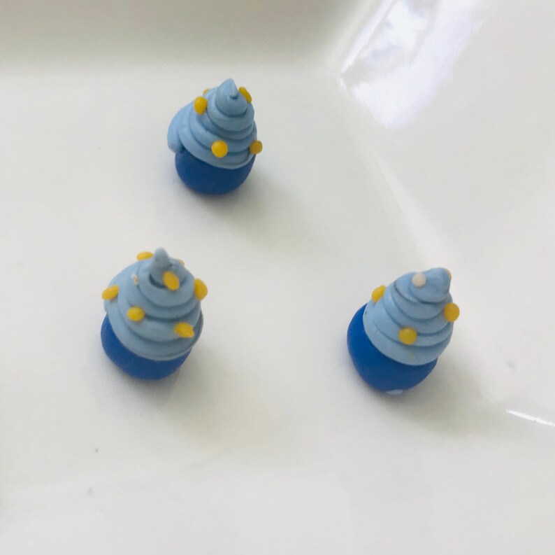 Pode incluir: Tr&ecirc;s mini cupcakes azuis e amarelos com cobertura branca. Os cupcakes s&atilde;o feitos de argila polim&eacute;rica e t&ecirc;m aproximadamente o tamanho de uma moeda de 10 c&ecirc;ntimos.