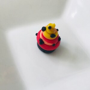 Pode incluir: Um pequeno cupcake vermelho, amarelo e preto com bolinhas pretas. O cupcake &eacute; decorado com redemoinhos de glac&ecirc; vermelho, amarelo e preto.