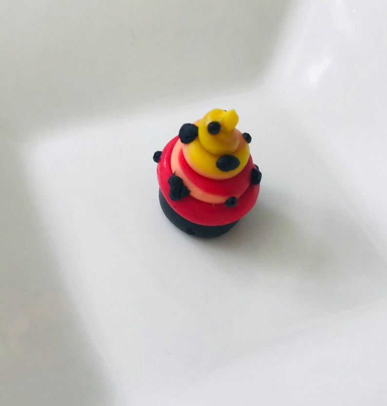 Pode incluir: Um pequeno cupcake vermelho, amarelo e preto com bolinhas pretas. O cupcake &eacute; decorado com redemoinhos de glac&ecirc; vermelho, amarelo e preto.