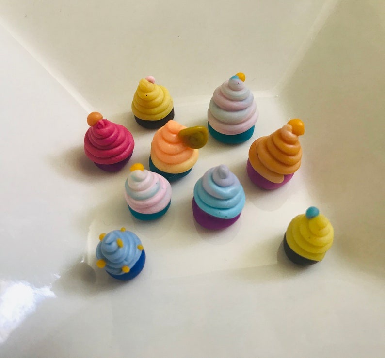 Pode incluir: Uma cole&ccedil;&atilde;o de dez mini cupcakes com cobertura colorida e granulado. Os cupcakes s&atilde;o feitos de argila polim&eacute;rica e est&atilde;o dispostos em um prato branco.