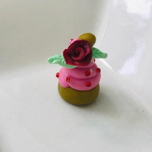 Pode incluir: Um pequeno cupcake rosa e verde com uma rosa vermelha no topo. O cupcake &eacute; decorado com granulado vermelho.