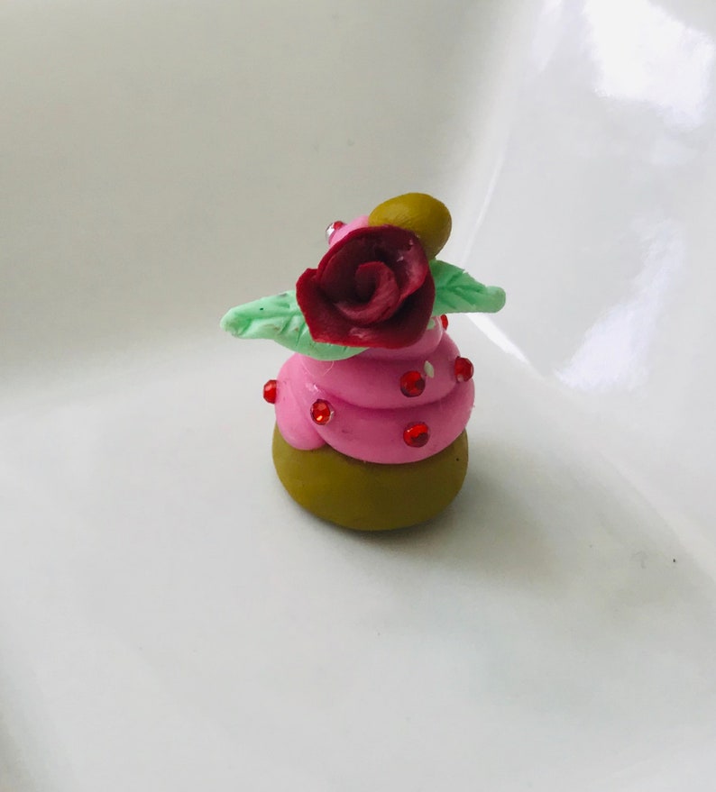Pode incluir: Um pequeno cupcake rosa e verde com uma rosa vermelha no topo. O cupcake &eacute; decorado com granulado vermelho.