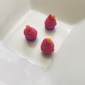 Pode incluir: Tr&ecirc;s mini cupcakes feitos de argila polim&eacute;rica. Os cupcakes s&atilde;o cor-de-rosa com fundos roxos e t&ecirc;m uma pequena bola laranja no topo.