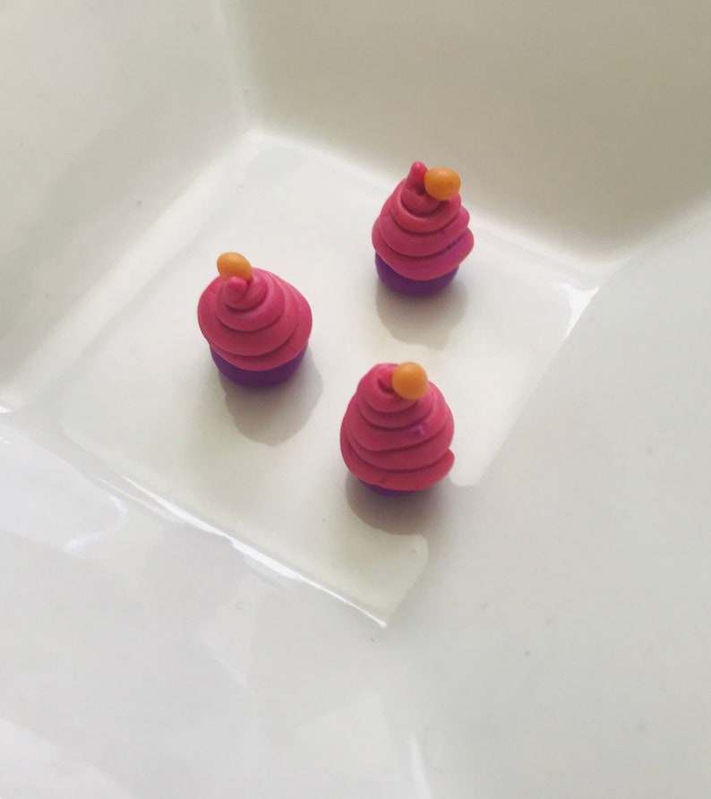 Pode incluir: Tr&ecirc;s mini cupcakes feitos de argila polim&eacute;rica. Os cupcakes s&atilde;o cor-de-rosa com fundos roxos e t&ecirc;m uma pequena bola laranja no topo.