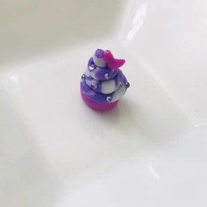 Pode incluir: Um pequeno cupcake em espiral roxo e branco com cobertura rosa e um cora&ccedil;&atilde;o rosa no topo. O cupcake &eacute; decorado com pequenas confeitas rosa e brancas.