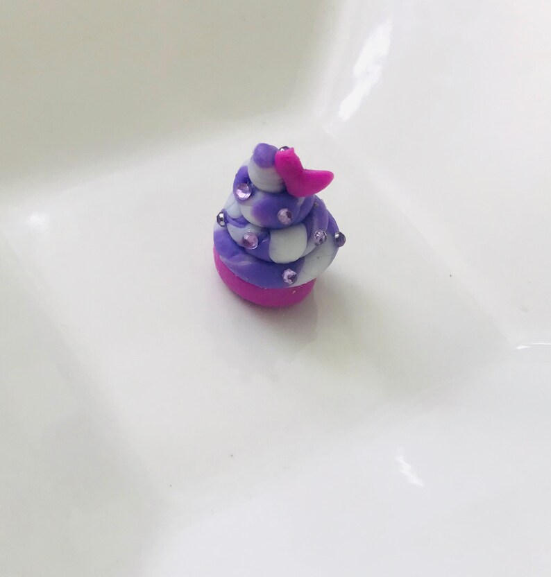 Pode incluir: Um pequeno cupcake em espiral roxo e branco com cobertura rosa e um cora&ccedil;&atilde;o rosa no topo. O cupcake &eacute; decorado com pequenas confeitas rosa e brancas.