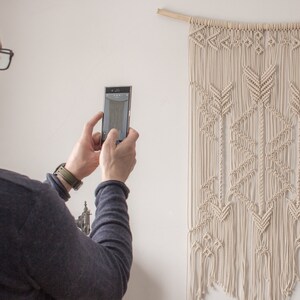 May include: A person is taking a photograph of a white macrame wall hanging with a geometric pattern of arrows. The wall hanging is made from natural cotton cord and is hung on a wooden dowel.
