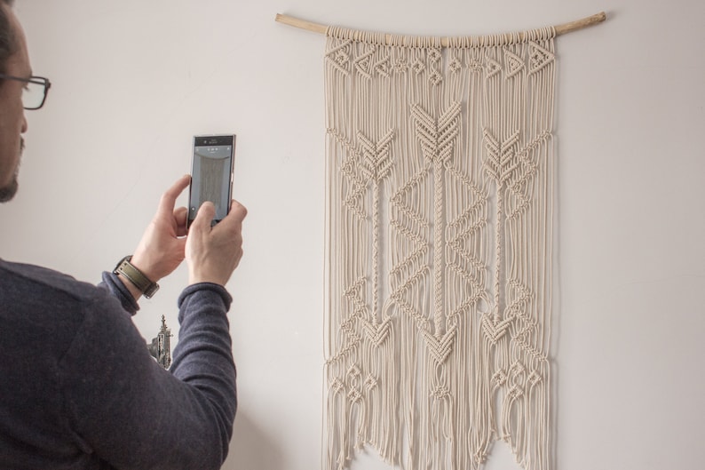May include: A person is taking a photograph of a white macrame wall hanging with a geometric pattern of arrows. The wall hanging is made from natural cotton cord and is hung on a wooden dowel.