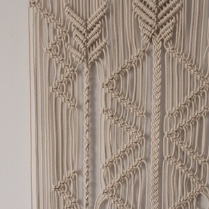 May include: A beige macrame wall hanging with a geometric pattern. The hanging is made of knotted cords and has a bohemian style.