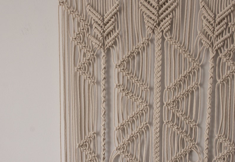 May include: A beige macrame wall hanging with a geometric pattern. The hanging is made of knotted cords and has a bohemian style.