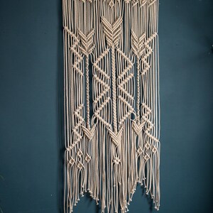 May include: A beige macrame wall hanging with a geometric pattern, featuring a wooden dowel at the top.