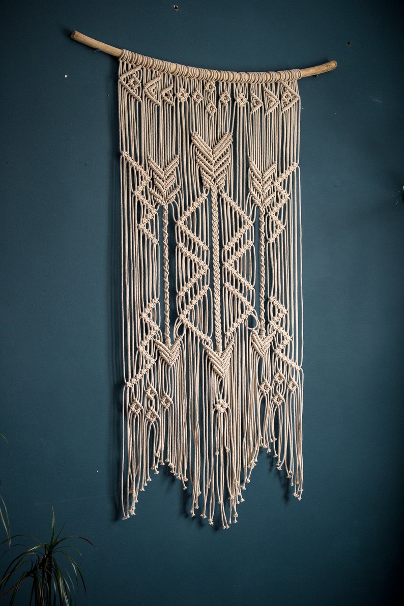 May include: A beige macrame wall hanging with a geometric pattern, featuring a wooden dowel at the top.