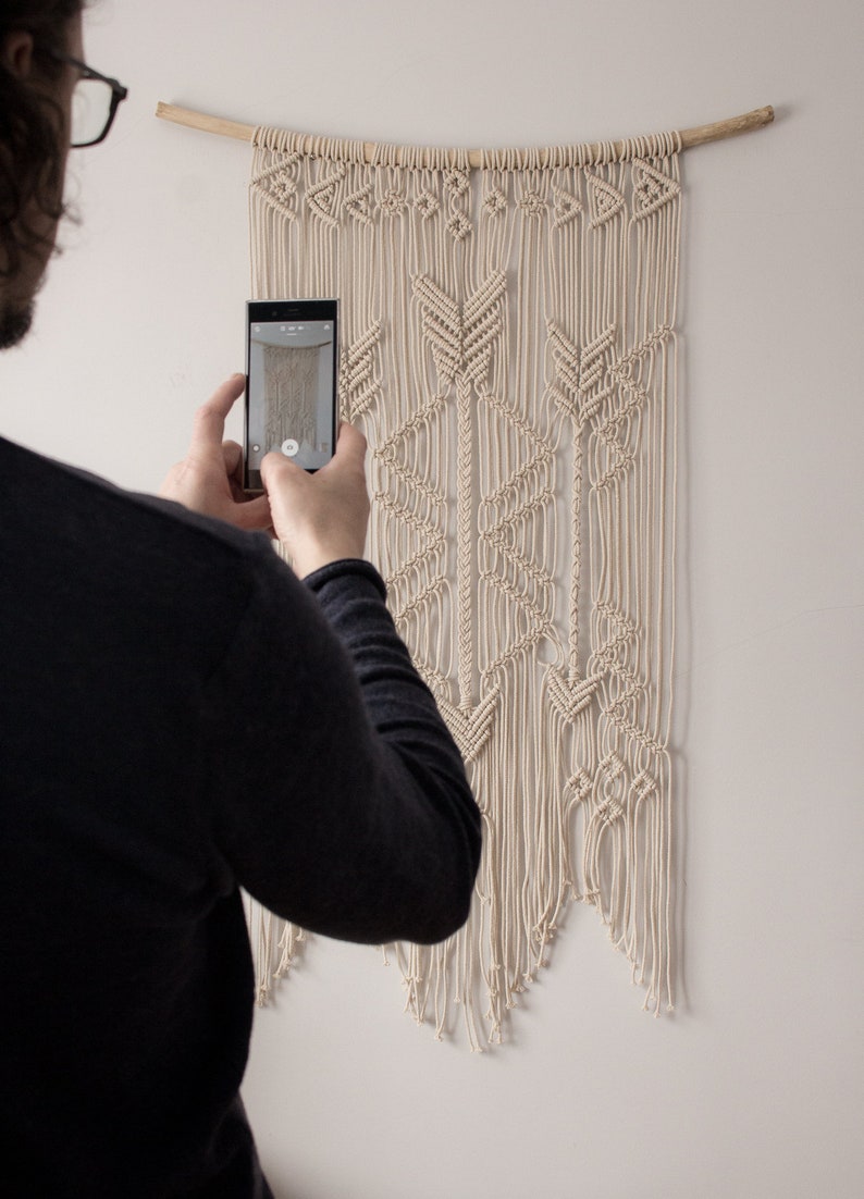 May include: A person taking a photograph of a macrame wall hanging with a geometric pattern. The wall hanging is made of natural coloured cotton cord and is hanging from a wooden dowel rod.