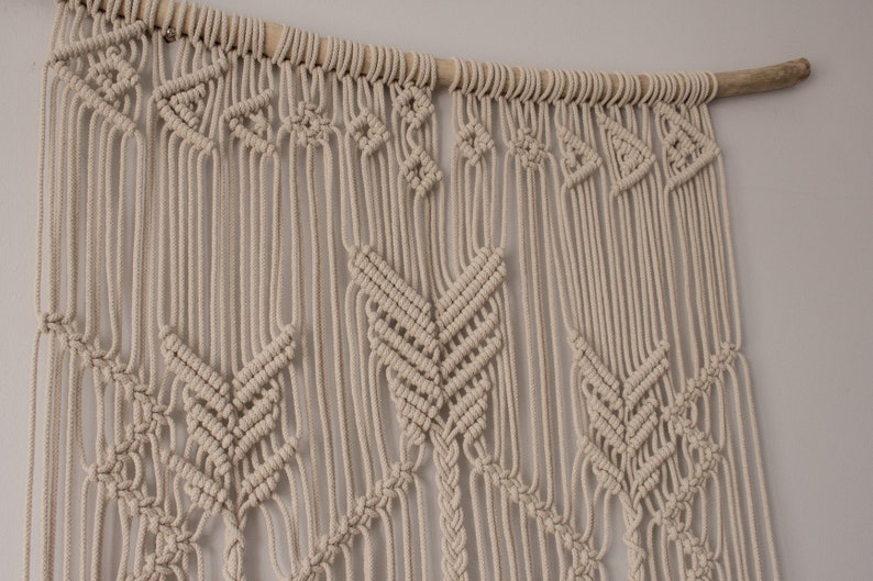 May include: A beige macrame wall hanging with a geometric pattern. The hanging is made of cotton cord and is attached to a wooden dowel.