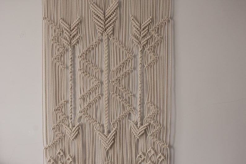 May include: A beige macrame wall hanging with a geometric pattern of arrows. The wall hanging is made of natural cotton cord and has a bohemian style.