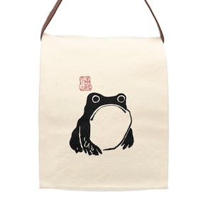 May include: A beige canvas tote bag with a brown leather strap. The bag features a black silhouette of a frog with a red stamp above it.