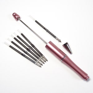 Könnte beinhalten: Ein zerlegter rosa Stift mit mehreren schwarzen Tintenminen. Die Stiftkomponenten umfassen eine Kappe, eine Spitze und eine Tintenpatrone. Der Stift hat eine metallisch rosa Farbe und ist zum Schreiben bestimmt. Die Nachfüllungen sind schwarz und haben klare Spitzen.