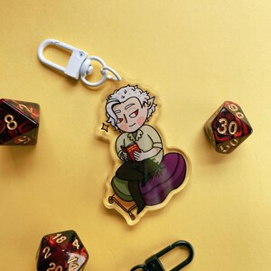May include: A white keychain with a cartoon illustration of a person with white hair and a book. The person is sitting on a purple cushion. The keychain is attached to a white metal ring. The background is yellow.