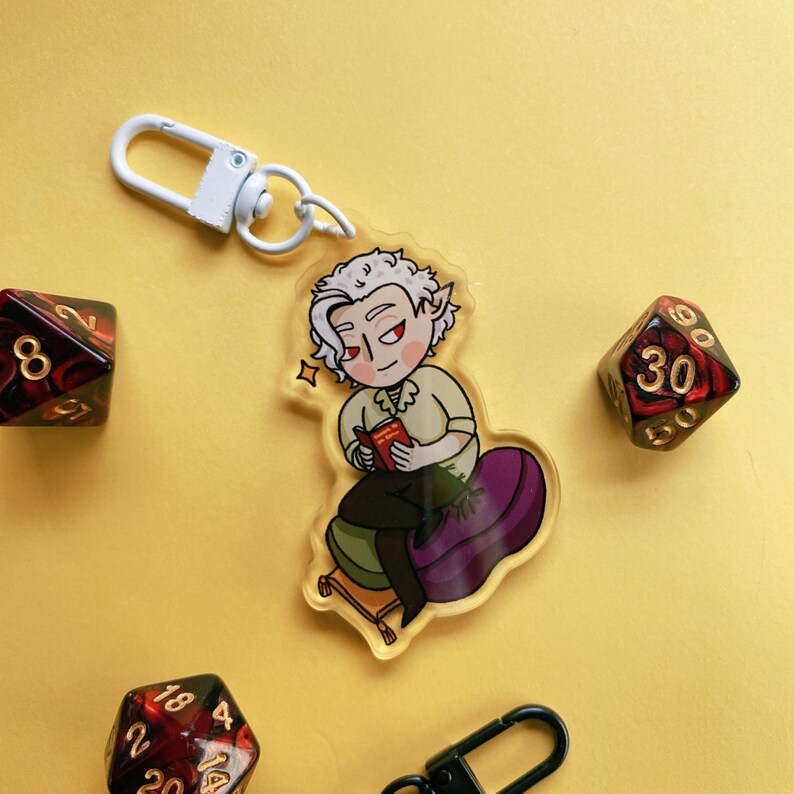 May include: A white keychain with a cartoon illustration of a person with white hair and a book. The person is sitting on a purple cushion. The keychain is attached to a white metal ring. The background is yellow.