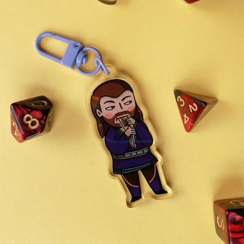May include: A purple and blue keychain with a cartoon character of a person with brown hair and a beard. The character is wearing a purple and gold outfit and holding a yellow object. The keychain is on a yellow background.
