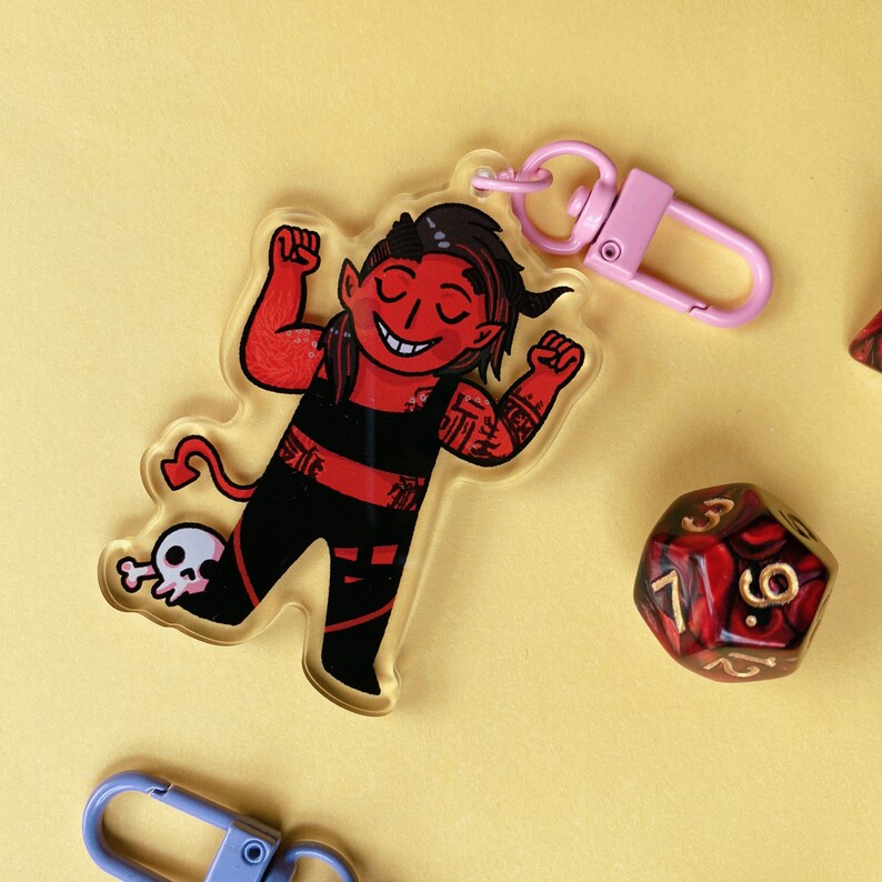 May include: Red and black cartoon devil character keychain with a skull and crossbones design. The keychain is attached to a pink metal clasp.