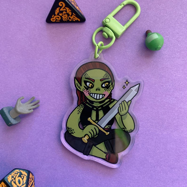 May include: A green and brown cartoon goblin character holding a silver sword. The character is sitting with their legs crossed and has a toothy grin. The character is on a clear acrylic keychain with a green metal clasp.