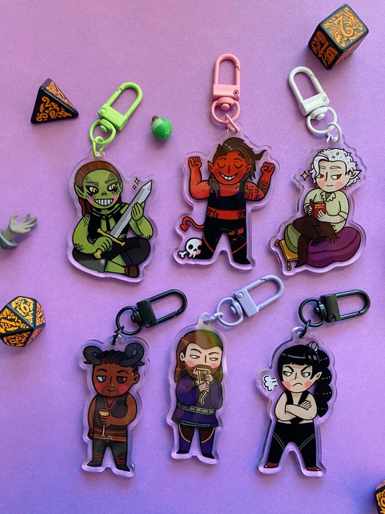 May include: Six colorful acrylic keychains featuring cartoon characters. Each keychain has a black metal ring and a silver or black metal clasp. The characters include a green goblin, a red demon, a white elf, a brown-skinned person with horns, a man with a beard, and a woman with black hair.