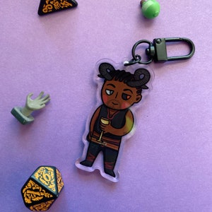 May include: A black and white cartoon character with horns and a goatee, holding a glass of wine. The character is printed on a clear acrylic keychain with a black metal ring.