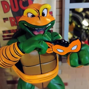Op de afbeelding: Een groene en oranje Teenage Mutant Ninja Turtle-actiefiguur met een ondeugende grijns, die een zwart en oranje masker draagt. De figuur houdt het masker in zijn hand.