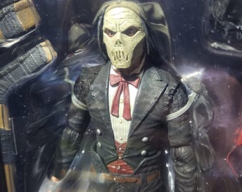 May include: A collectible action figure featuring a character with a pale, skull-like mask, wearing a black textured suit jacket, a white shirt, a red bow tie, and a red vest. The figure is encased in clear plastic packaging with visible joints.
