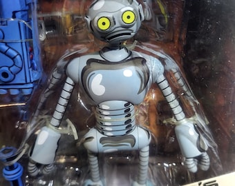May include: A gray robot action figure with large yellow eyes and a segmented torso, packaged in a clear plastic box. The packaging features the text Eastman and Laird's Teenage Mutant Ninja Turtles at the bottom with a blue accessory included.